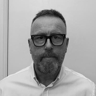 A man with glasses and a beard wearing a white shirt against a plain background.