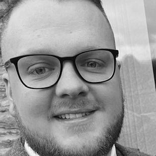 A person with glasses smiling in front of a window, wearing a suit and shirt.